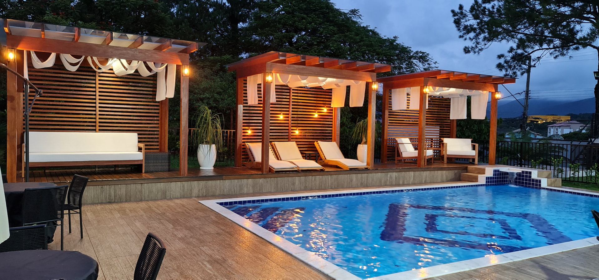 Área de lazer com piscina, pergolados iluminados e deck de madeira ao anoitecer